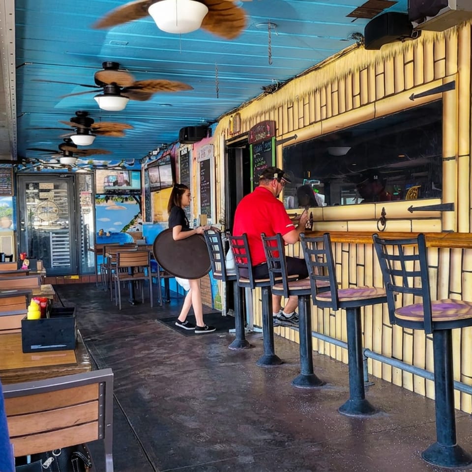 Casual restaurant interior with bamboo-style walls, blue ceiling, bar seating, ceiling fans, and diners at tables.