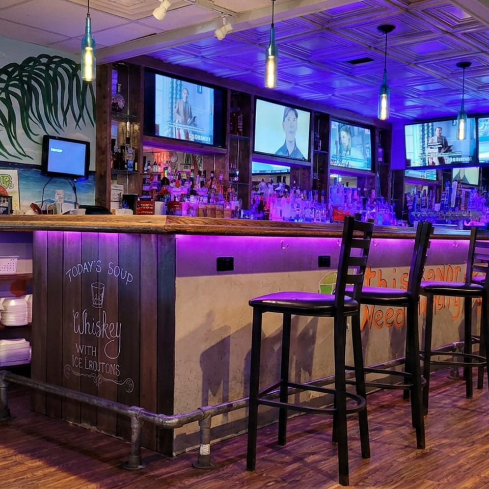 Vibrantly lit tropical-themed restaurant bar with purple lighting, high stools, multiple televisions, and colorful murals.