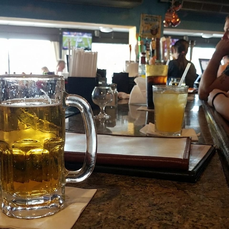 Pint of golden beer on a bar beside a yellow cocktail, menus, and patrons in a casual restaurant.