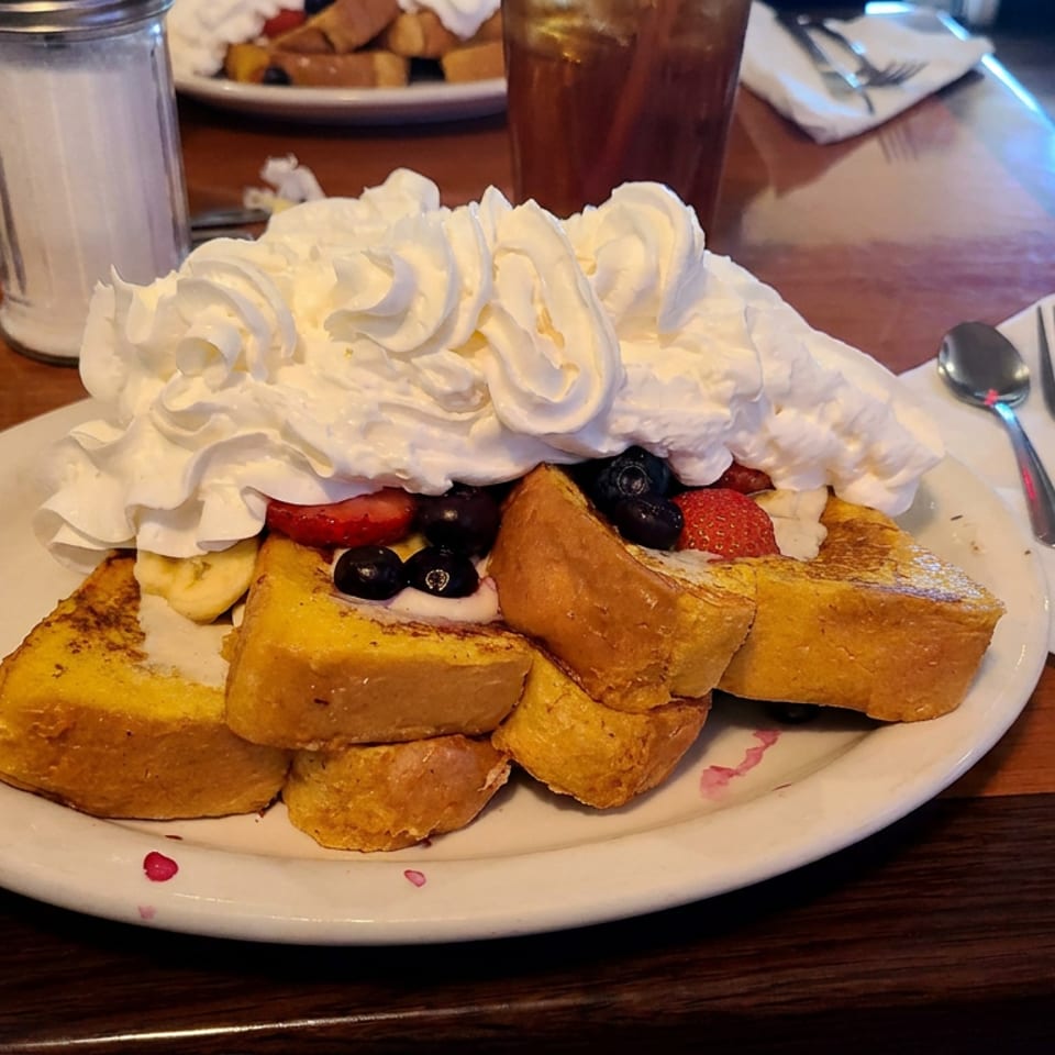 Plate of golden French toast topped with whipped cream, strawberries, bananas, and blueberries at a restaurant table.