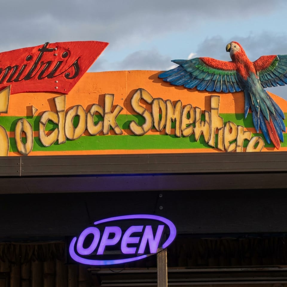 Colorful Tiki restaurant facade features a parrot, tropical sign, and glowing purple open sign.