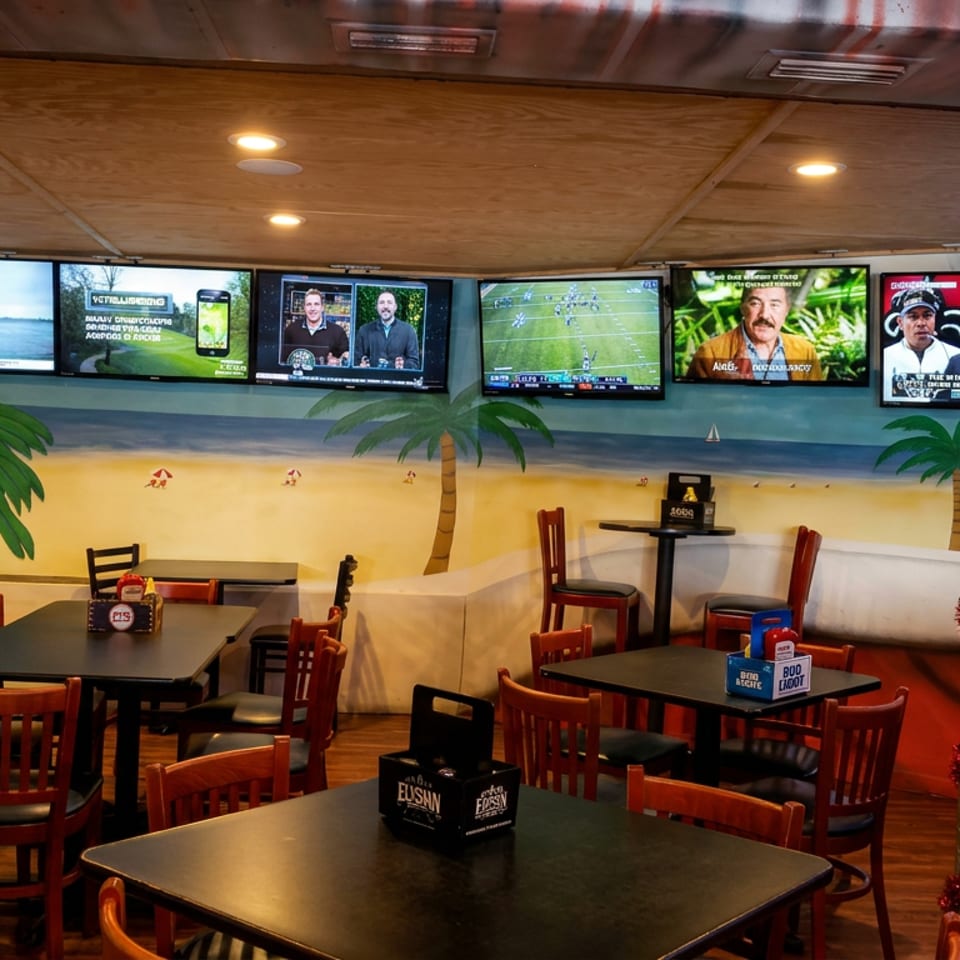 Sports bar dining area with empty tables, beach mural, palm trees, and multiple televisions showing sports and news.