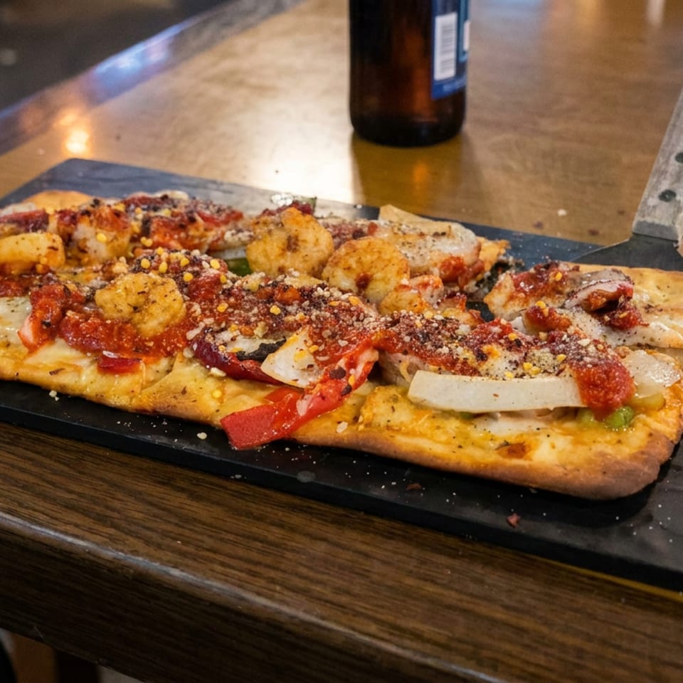 Rectangular pizza topped with shrimp, tomatoes, peppers, onions, cheese, and herbs served on a black slate board.